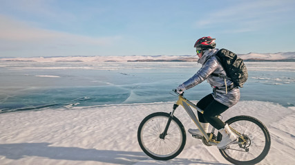 Woman is riding bicycle on the ice. Girl is dressed in a silvery down jacket, cycling backpack and...