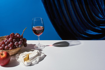 classic still life with fruits, red wine and Camembert cheese on white table near velour curtain...