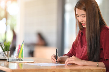 Portrait of beautiful woman looking good in red shirt while writing on the paper and sitting at the...