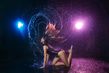A slender brunette girl in a swimsuit, waves her head so that water flows from her wet hair, on a black background in the studio under raindrops, backlighting in blue and pink spotlights.