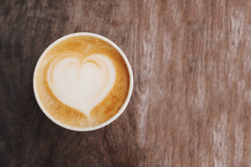 Paper cup of cappuccino coffee with latte art in the shape of heart on wooden background.