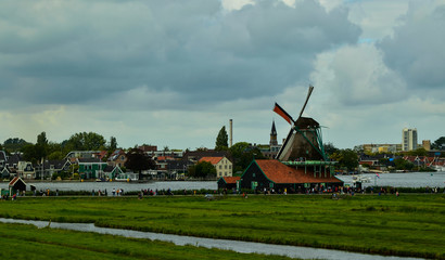 Fototapeta premium Zaanse Schans, Holland, August 2019. North-east of Amsterdam is a small community located on the quay of the Zaan river. View from above of the fields with mills, tourists are noticed. Cloudy day.