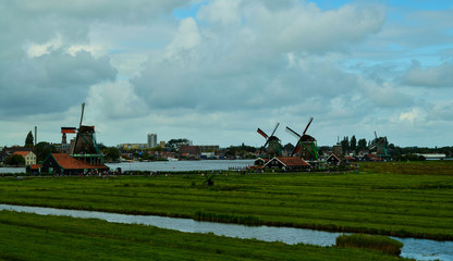 Fototapeta premium Zaanse Schans, Holland, August 2019. North-east of Amsterdam is a small community located on the quay of the Zaan river. View from above of the fields with mills, tourists are noticed. Cloudy day.