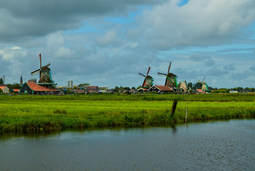 Fototapeta premium Zaanse Schans, Holland, August 2019. North-east of Amsterdam is a small community located on the quay of the Zaan river. View from above of the fields with mills, tourists are noticed. Cloudy day.