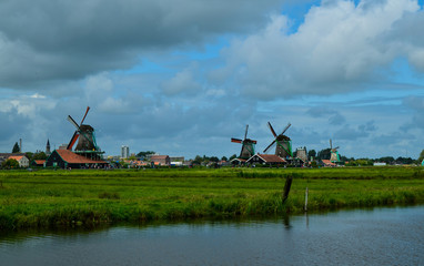 Fototapeta premium Zaanse Schans, Holland, August 2019. North-east of Amsterdam is a small community located on the quay of the Zaan river. View from above of the fields with mills, tourists are noticed. Cloudy day.