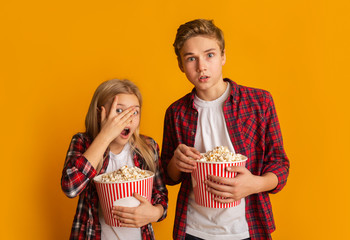 Scared brother and sister holding popcorn buckets and looking at camera