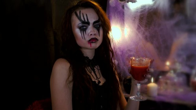 Girl with makeup for Halloween is sitting in front of a mirror. Woman drinks a red drink from a glass and shows her middle finger.