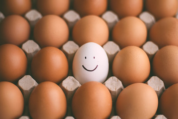 Many brown eggs and one white smiling egg in the middle of carton background.