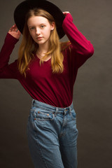 Studio portrait of young blonde girl in a black hat, red blouse and blue jeans on a brown background.