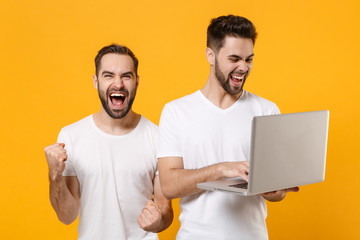 Joyful young men guys friends in white t-shirts isolated on yellow orange background studio portrait. People lifestyle concept. Mock up copy space. Working on laptop pc computer doing winner gesture.