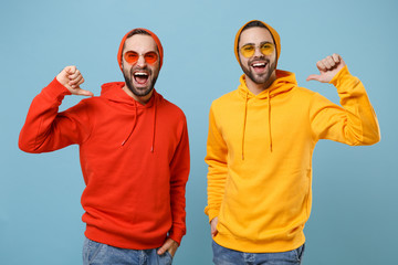 Two cheerful hipster men guys in fashion red yellow clothes eyeglasses posing isolated on pastel blue background in studio. People lifestyle concept. Mock up copy space. Pointing thumbs on themselves.