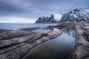 View over Ersfjord from colorful rocks and rockpools to snowy Oksen mountains on a dark cloudy day in Winter, Cape Tungeneset Senja Norway