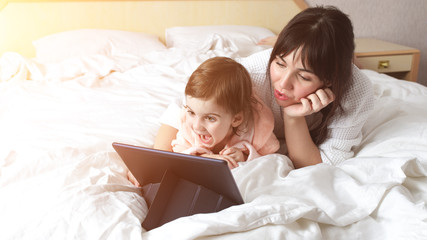 Little baby girl and her mother are lying on the bed and watching on tablet pc and daughter is laughing. Happy family spending time together. Speaking with father by video communication call.