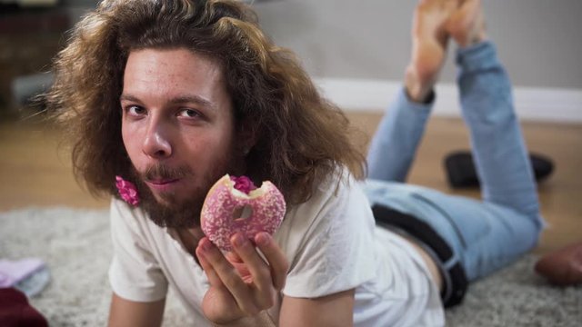 Close-up of positive Caucasian intersex person lying on the floor and eating tasty donut. Happy male woman with brown eyes and flowers in beard looking at camera and smiling.