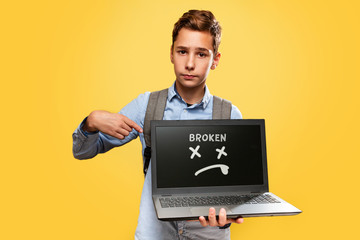 Concept of modern technologies and education. A disgruntled Caucasian teenage boy holds an open laptop and points at it with his finger. Yellow background. Copy space. The inscription Broken