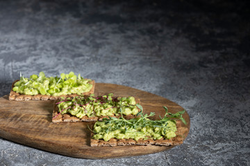 Three toast on a rye cracker with tender avocado and sprouts of micro green beetroot pea salad on a wooden Board and a gray concrete background.