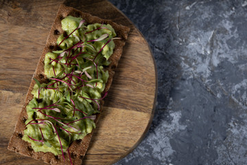Toast on a rye cracker with tender avocado and sprouts of micro beet greens on a wooden plank and a gray concrete background. Top view