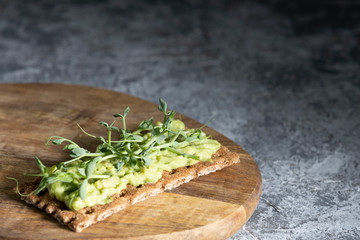 Healthy vegitarian Breakfast. Toast on a rye cracker with tender avocado and sprouts of micro peas greens on a wooden plank and a gray concrete background.
