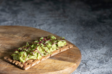 Healthy vegitarian Breakfast. Toast on a rye cracker with tender avocado and sprouts of micro beet greens on a wooden plank and a gray concrete background.