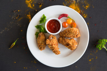  Roasted breaded chicken on a white plate and ketchup sauce. On the table are cherry tomatoes, chili peppers, parsley and sprinkled with powder of various spices.