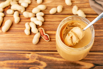 Creamy peanut paste in open glass jar, peanut butter in spoon. Peanuts in the peel scattered on the brown wooden table with copy space for cooking breakfast. Vegan food concept.