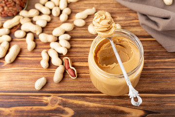 Creamy peanut paste in open glass jar, peanut butter in spoon. Peanuts in the peel scattered on the brown wooden table with copy space for cooking breakfast. Vegan food concept.