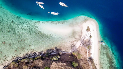 Top down drone shot of a paradise island with some boats anchored around in Komodo National Park, Flores, Indonesia. Brownish island turns into white sand beach and further into turquoise and navy sea