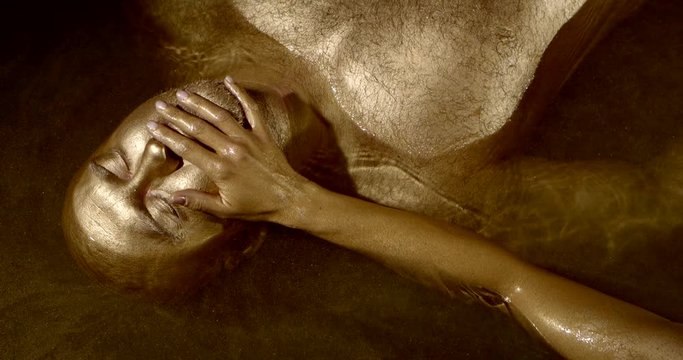 man is lying in water tub, his body and face covered by gold paint, female hand is touching him