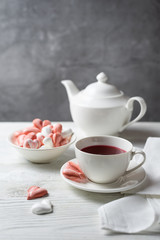 Beautiful romantic breakfast with red tea Hibiscus and meringue cookies in the shape of hearts and candy. White tea-set stand on a wooden table with a concrete background. Vertical with copy space.