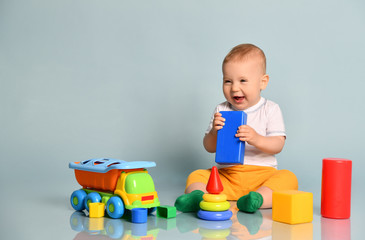 Cute smiling laughing infant baby boy toddler in yellow pants is sitting surrounded by toys, holding block at his mouth