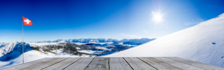 wooden shelf display table top against blue winter mountain panorama and swiss flag snow covered blue mountain layers