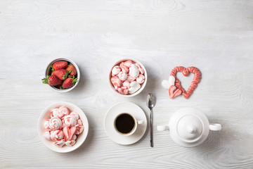 Beautiful romantic breakfast coffee with cream, strawberries and meringue cookies in the shape of hearts. White set stands in a row on a wooden table. Flat lay of food knolling with copy space.