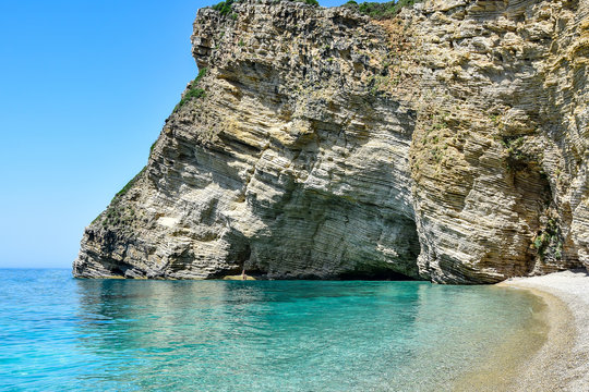 Paleokastritsa / Corfu, Greece - Chomi beach (Paradise), white-yellow-brown rocks in layers, a white pebble beach near the transparent turquoise Ionian Sea, in the summer afternoon