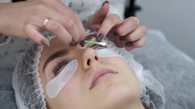 An esthetician is applying the lash adhesive equally on the lashes. The lash artist is working in a disposable mask. The client is lying on a couch in a disposable hat.