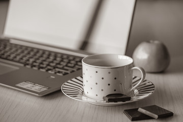 Cup of coffee on the wooden table with notebook on the background. Piece of black chocolate is on the saucer, break time in the office for refreshment