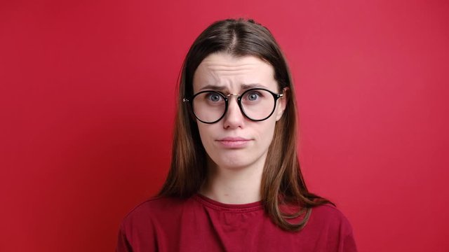  Resentful girl in glasses blowing his cheeks while being offended at somebody showing his negative emotions and resentment, wears  t-shirt, stands over red background 
