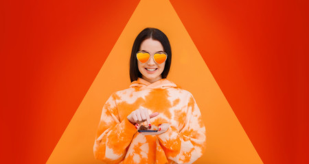 Portrait of beautiful woman in heart-shaped sunglasses. Pretty girl in hoody with smartphone in hands addicted to social media. Bright geometric trendy background. Free space for text. Trends 2020.