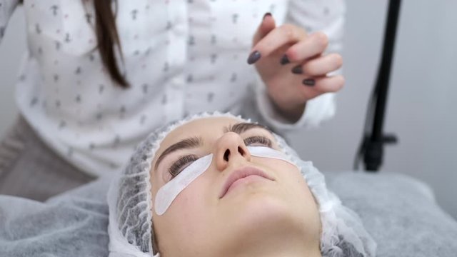 An esthetician is applying the lash adhesive equally on the lashes. The lash artist is working in a disposable mask. The client is lying on a couch in a disposable hat.