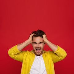 crazy man in yellow shirt holding head and screaming isolated on red background