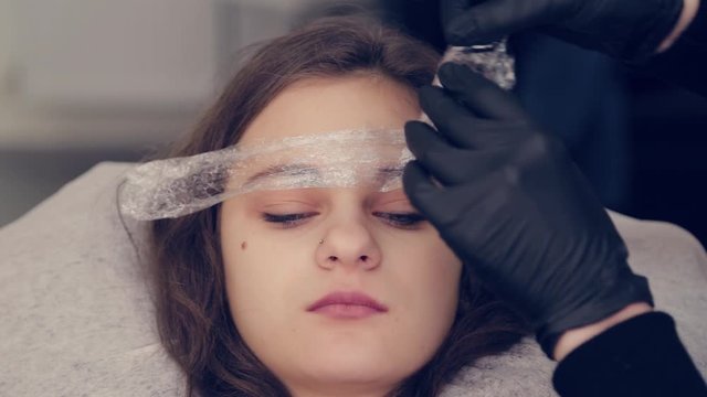 Professional master eyebrow woman removes the film from the eyebrow and removes liquid on the eyebrows in a beauty salon.