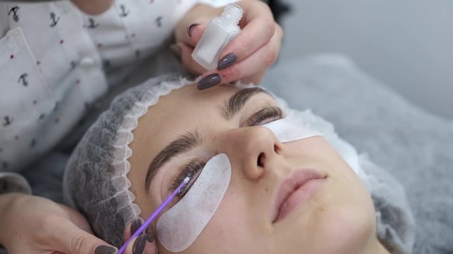 A lash artist is applying the lash adhesive equally on the lashes. The lash maker is working in a disposable mask. The client is lying on a couch in a disposable hat.