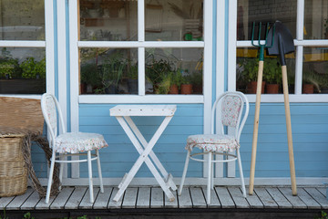 Rustic porch with white wooden table with chairs and garden tools near wooden blue house in countryside.