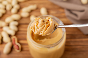 Peanut butter in spoon near creamy peanut paste in open glass jar. Peanuts in the peel scattered on the brown wooden table with copy space for cooking breakfast. Vegan food concept.