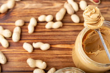 Creamy peanut paste in open glass jar, peanut butter in spoon. Peanuts in the peel scattered on the brown wooden table with copy space for cooking breakfast. Vegan food concept.
