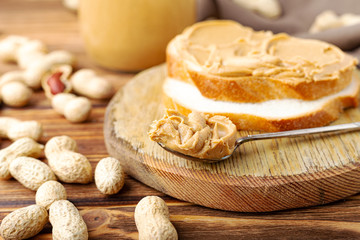 Peanut butter in spoon near creamy peanut paste in open glass jar, slice of peanut butter bread, toast. Peanuts in the peel scattered on the brown wooden table with copy space for cooking breakfast