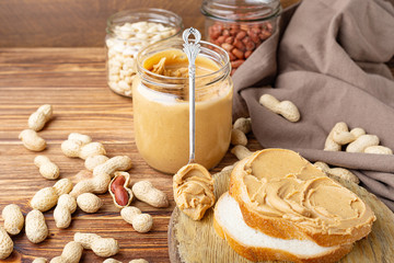 Peanut butter in spoon near creamy peanut paste in open glass jar, slice of peanut butter bread,...