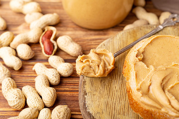 Peanut butter in spoon near creamy peanut paste in open glass jar, slice of peanut butter bread,...