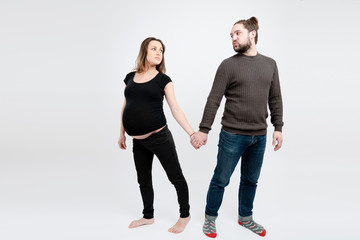 Young attractive couple in casual clothes, bearded man and pregnant woman standing together, holding hand in hand and looking at each other, isolated over white background
