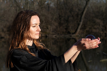 Woman with dark long hair in black robes is holding polished Shungite Sphere in her hands in front...