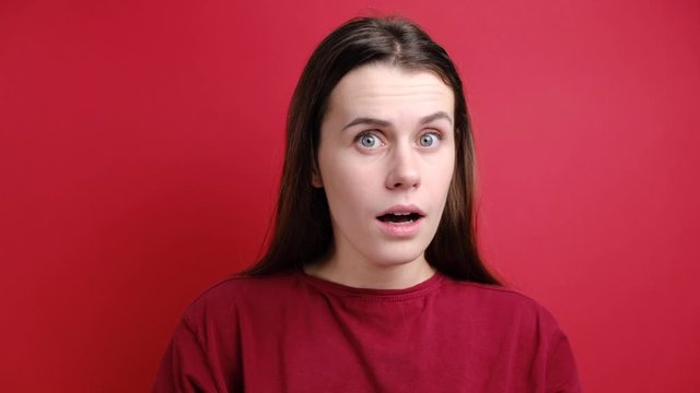 Pretty young woman stares at camera with shocked expression, recieves unexpected news, standing isolated over red background. Positive human emotion. Great surprisment or unexpectedness